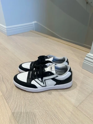 Svarta och vita Vans sneakers - Snygga svartvita sneakers från Vans med klassisk låg siluett. Skorna har svart mocka och vitt läder, perforerad tå och svart snörning. Den ikoniska Vans-loggan syns på sidan och yttersulan är vit med brun undersida. Använda fåtal gånger
