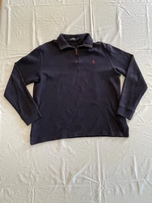 Mörkblå quarter zip från Polo Ralph Lauren - Mörkblå långärmad quarter zip-tröja från Polo Ralph Lauren med klassisk krage och dragkedja med läderdetalj. Broderad röd logga på bröstet. Perfekt för en stilren och avslappnad look.     Tveka inte, hör av er vid intresse, frågor eller prisförslag!