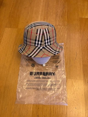 Burberry bucket hat  - Snygg bucket hat från Burberry i beige med det ikoniska rutmönstret i svart, vitt och rött. Hatten har rund form och metallöljetter på sidorna för ventilation. Perfekt accessoar för en trendig streetstyle-look.