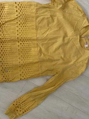 Gul blus med broderade detaljer - Snygg gul blus från Odd Molly med långärmad design. Blusen har broderade och hålmönstrade detaljer på nedre delen och ärmsluten, vilket ger en unik och trendig look. Tillverkad i ett lätt och luftigt material som passar perfekt till vår och sommar.