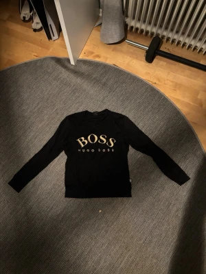 Svart långärmad tröja Hugo Boss - Svart långärmad tröja från Hugo Boss med stort guldigt BOSS-tryck på bröstet. Klassisk rund halsringning och stilren design, perfekt för dig som gillar snygga logoplagg. Storlek XS men passar S