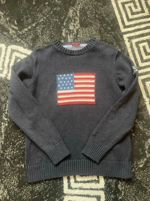 Mörkblå stickad tröja från Gant - Snygg mörkblå stickad tröja från Gant med amerikansk flagga på bröstet och broderad text 'GANT NEW HAVEN 1949' på ärmen. Tröjan har rund halsringning, långa ärmar och ribbade muddar. Perfekt för en cool och avslappnad stil.