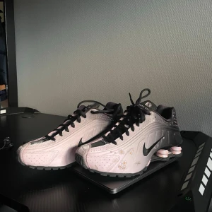 Nike Shox rosa och svarta sneakers - Nike Shox sneakers i en snygg kombination av ljusrosa och svart med silvriga detaljer. Skorna har den ikoniska Shox-sulan med fjädrande dämpning och svart logga på sidan. Ovandelen är i syntetmaterial med perforeringar och dekorativa sömmar.