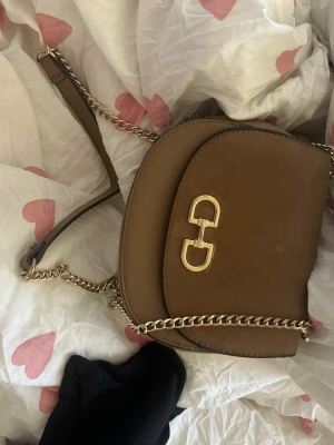 Brun axelväska från Salvatore Ferragamo - Snygg brun axelväska i läder från Salvatore Ferragamo med guldfärgad kedja och ikoniskt metallspänne framtill. Väskan har rundad form och stilren design, perfekt för att lyfta din outfit.