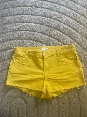 Gula jeansshorts med fransig kant - Snygga gula jeansshorts med fransig kant och klassisk femficksdesign. Shortsen har bälteshällor och stängs med knapp och dragkedja. Perfekta för sommardagar när du vill sticka ut lite extra.