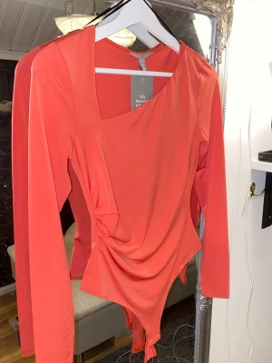 Korallröd asymmetrisk body H&M - Trendig korallröd body från H&M med lång ärm och asymmetrisk halsringning. Snygg rynkdetalj på sidan och tillverkad i stretchigt material med 93% återvunnen polyester. Perfekt för dig som vill sticka ut med en unik look.