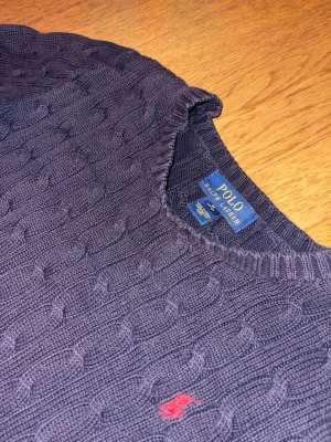 Mörkblå kabelstickad tröja Polo Ralph Lauren - Kabelstickad tröja i mörklila från Polo Ralph Lauren med rund halsringning och klassisk röd broderad logga på bröstet. Tröjan har långa ärmar och ribbade muddar vid ärmslut och nederkant. Perfekt för en stilren och avslappnad look. Tröjan har två st färgfläckar på sig som är synliga på bilden som ger en liten schysst look med tanke på att den matchar med färgen på loggan. Tröjan passar en kille som är ungefär 160 cm lång. 