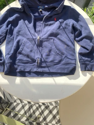 Marinblå hoodie från Polo Ralph Lauren - Snygg marinblå hoodie från Polo Ralph Lauren med dragkedja och klassisk röd logga på bröstet. Hoodien har känguruficka framtill och är tillverkad i mjukt bomullsmaterial. Perfekt för en avslappnad och stilren look.