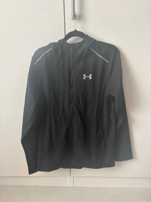 Svart vindjacka från Under Armour - Svart vindjacka från Under Armour med huva och dragkedja framtill. Jackan har vita detaljer på axlarna och logga på bröstet samt stor logotyp på ryggen. Lätt material som passar perfekt för blåsiga dagar.