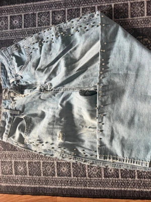Ljusblå jeans med nitar och slitningar - Säljer ett par ljusblå jeans med coola slitningar och nitar längs sidorna. Jeansen har en relaxed fit och hög midja, perfekta för en trendig och avslappnad stil. Materialet är klassisk denim i bomull och de har dragkedjegylf.