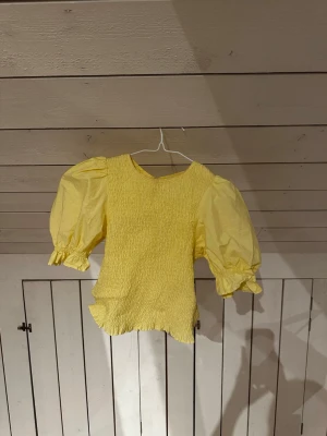 Gul puffärmad topp med smock - Gul söt topp, figur formad. Har en svag defekt på inner sidan (kom priv för bild) syns knappast och går förmodligen bort i en tvätt.