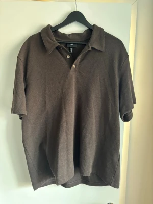 Brun pikétröja från H&M Regular Fit - Brun pikétröja från H&M i regular fit med klassisk krage och knappar framtill. Tröjan har korta ärmar och är tillverkad i ett mjukt bomullsmaterial. Perfekt för en avslappnad och stilren look.