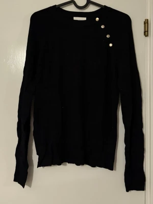 Svart ribstickad tröja från H&M - Svart stickad tröja från H&M Basics med rund halsringning och dekorativa silverfärgade knappar på ena axeln. Långärmad modell i mjukt material, perfekt för lager-på-lager och enkel att matcha med jeans eller kjol.