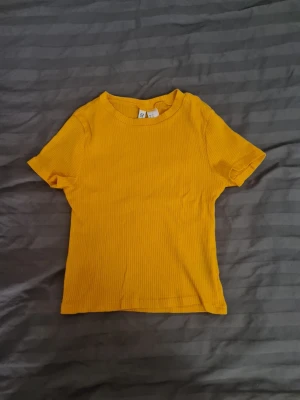 Gul ribbad t-shirt från H&M Divided - Snygg gul ribbad t-shirt från H&M Divided i modellen Basic. T-shirten har korta ärmar, rund halsringning och är tillverkad i ett stretchigt, mjukt material som sitter skönt på kroppen. Perfekt för en färgglad och trendig look.