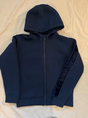 Marinblå hoodie från Emporio Armani - Snygg marinblå hoodie från Emporio Armani med dragkedja framtill och huva. På ena ärmen finns en stilren sammetsdetalj med märkesloggan broderad. Perfekt för dig som gillar exklusiva streetwear-plagg.
