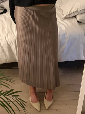 Brun långkjol från Vila - Snygg siden långkjol från Vila i en mjuk brun nyans. Kjolen har ett brett midjeband och ett glansigt, flowy tyg som ger en lyxig känsla. Perfekt för dig som gillar stilrena och trendiga plagg.