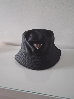 Svart bucket hat från Prada - Snygg svart bucket hat från Prada i glansigt nylonmaterial. Klassisk form med bred, nedåtlutande brätte och ikoniska metalltriangeln med logga framtill. Perfekt accessoar för en trendig streetwear-look.