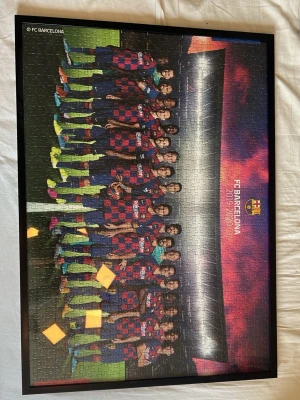 FC Barcelona pussel i ram - Säljer ett pussel med motiv av FC Barcelona-laget, monterat i en svart ram. Pusslet har färgstarka detaljer med spelare i blå-röda matchställ och bakgrund i lila och rosa toner. Perfekt för dig som älskar fotboll och vill ha en unik väggdekoration.
