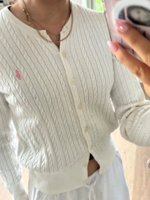 Vit kabelstickad kofta från Ralph Lauren - Vit kabelstickad kofta från Ralph Lauren med rund halsringning och knäppning framtill. Liten rosa broderad logga på bröstet. Mjuk och skön bomullskvalitet, perfekt för lager-på-lager. Klassisk och tidlös design.