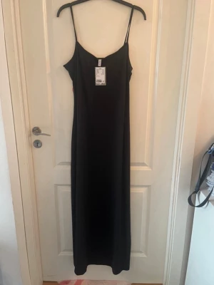 Svart långklänning med tunna band - En stilren svart långklänning med tunna axelband och rak passform. Klänningen är tillverkad av återvunnen polyester och har en minimalistisk design som passar perfekt till många olika looks.