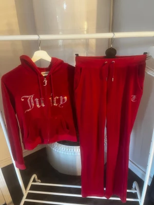 Rött Juicy Couture velourset - Säljer ett ikoniskt rött velourset från Juicy Couture med hoodie och byxor. Hoodien har dragkedja, fickor och glittrande Juicy-tryck framtill. Byxorna har dragsko i midjan, fickor och Juicy-tryck i strass på baksidan. Perfekt för en chill och trendig look. Byxorna är storlek XS och tröjan M. Båda för 1000kr eller enskilt för 600kr. Använt väldigt få gånger.