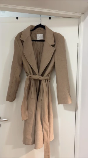 Beige lång kappa med skärp - Stilren beige kappa med klassisk krage, knytskärp i midjan och dubbelknäppning framtill. Kappan har två sidofickor och är helfodrad för extra komfort. Perfekt för dig som vill ha en tidlös och snygg look till hösten eller vintern.