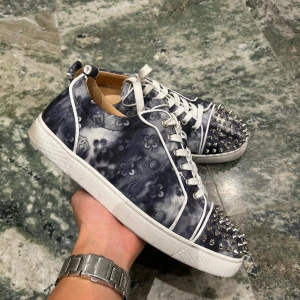 Christian Louboutin sneakers med nitar - Unika sneakers från Christian Louboutin med blått och vitt batikmönster, silverfärgade nitar på tån och klassisk röd sula. Skorna har vita snören och detaljer i vitt längs kanterna. Perfekt för dig som vill sticka ut med en edgy och lyxig stil.