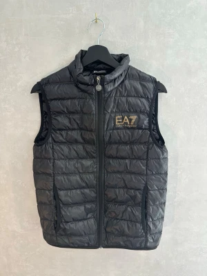 Svart dunväst från EA7 Emporio Armani - Snygg svart dunväst från EA7 Emporio Armani med quiltad design och hög krage. Västen har dragkedja framtill, två sidofickor och en guldfärgad logga på bröstet. Tillverkad i lätt och glansigt material som ger en sportig look.