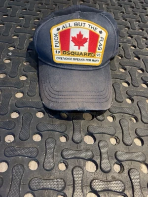 Mörkblå Dsquared2 keps med patch - Säljer en mörkblå keps från Dsquared2 med stor patch framtill med kanadensisk flagga och text. Justerbar rem bak och broderad gul text. Klassisk böjd skärm och tillverkad i bomullstyg. Perfekt för dig som gillar streetwear och statement-plagg.