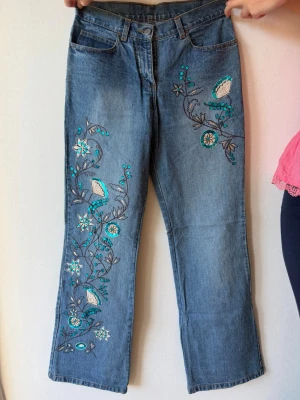 Blå broderade jeans Benetton - Snygga blå jeans från United Colors of Benetton med unika broderier i turkos, vitt och grått längs ena benet. Rak passform och klassisk femficksmodell. Perfekta för dig som vill sticka ut med coola detaljer och färgglada inslag.