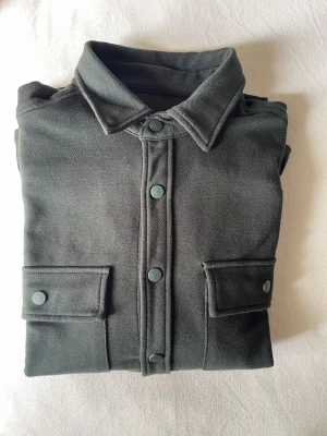 Mörkgrön overshirt med tryckknappar - Stilren mörkgrön overshirt med klassisk krage och tryckknappar framtill. Två stora bröstfickor med lock och knappdetaljer. Overshirten har en rak passform och är tillverkad i ett mjukt, tjockare tyg som ger en avslappnad look.