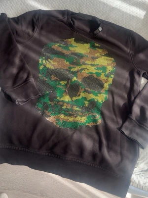 Svart Philipp Plein hoodie med döskalle - Svart hoodie från Philipp Plein med en stor döskalle framtill i camouflagemönster i grönt, gult och brunt. Klassisk passform med ribbade muddar och rund hals. Perfekt för dig som vill sticka ut med en edgy streetstyle. Står Xl men tvättade den fel så de blev som en S eller M jag är 173 och väger 80kg o den sitter exakt som ja vill rätt så tajt men bra längd 