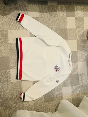 Vit Moncler sweatshirt med logga - Vit sweatshirt från Moncler med broderad logga på bröstet och ärmen. Tröjan har långa ärmar och ribbade muddar samt nederkant i rött och marinblått. Klassisk och sportig look med rund halsringning.