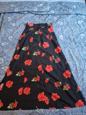 Svart långkjol med röda rosor - Svart långkjol med vackra rosor. Den utsvängda silhuetten ger ett elegant och romantiskt fall. Inköpt i vintage butik