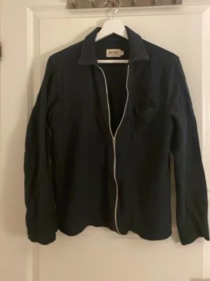 Svart overshirt med dragkedja - Stilren svart overshirt med vit dragkedja framtill och klassisk krage. Jackan har lång ärm och en diskret bröstficka. Perfekt för lager-på-lager och enkel att matcha med olika outfits.