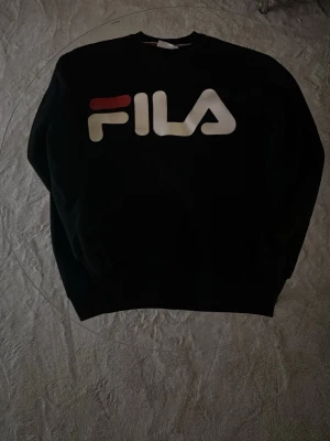 Svart sweatshirt från FILA - Svart sweatshirt från FILA med stor logga i vitt och rött framtill. Klassisk rund halsringning och ribbade muddar vid ärmslut och nederkant. Perfekt för en sportig och avslappnad stil.