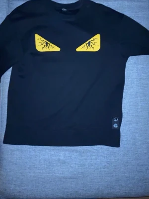 Svart Fendi tröja med gula ögon - Säljer en svart tröja från Fendi med ikoniska gula ögon framtill. Tröjan har rund halsringning och är tillverkad i ett mjukt material. Perfekt för dig som vill sticka ut med en unik designerstil. 