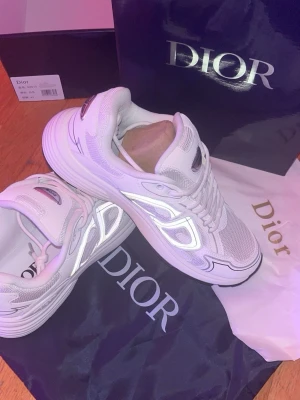 Vita Dior sneakers med reflekterande detaljer - Säljer ett par vita Dior sneakers med reflekterande detaljer och meshpaneler. Skorna har en chunky sula, klassisk snörning och logga på plösen. Perfekta för dig som vill ha en clean och exklusiv look med futuristiska vibbar.