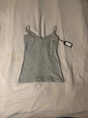 Grå ribbad linnetopp - Snygg grå linnetopp med smala axelband och v-ringad hals. Toppen är ribbad och har en enkel, stilren design som passar perfekt till jeans eller kjol. Materialet känns mjukt och stretchigt, vilket gör den bekväm att bära.