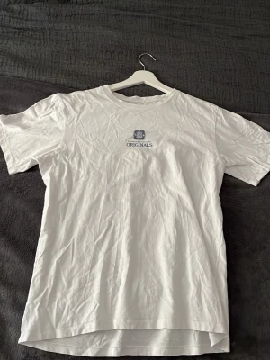Vit t-shirt med stor grafisk print - Säljer en vit t-shirt med rund hals och korta ärmar. På bröstet finns en liten logga med texten 'ORIGINALS' och på ryggen en stor, detaljerad grafisk print i svart och grått med texten 'LIMITED EDITION'. Perfekt för dig som gillar stilrena men ändå unika plagg.