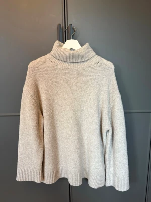 Beige polotröja i stickat material - Mysig beige stickad polotröja med bred ribbad krage och muddar. Tröjan har en avslappnad passform och är tillverkad i ett mjukt material som passar perfekt för kyliga dagar. Enkel och stilren design som är lätt att matcha.