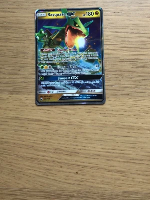 Rayquaza GX Pokémonkort - Säljer ett Rayquaza GX Pokémonkort med 180 HP. Kortet har en färgstark design med Rayquaza i grönt och gult mot en blå bakgrund. Det är ett samlarkort i standardstorlek med holografiska detaljer och engelskt texttryck.