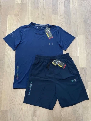 Blått träningsset från Under Armour - Snyggt träningsset från Under Armour med en blå t-shirt och matchande shorts. T-shirten har diskret logga på bröstet och text längs sidan. Shortsen har resårmidja med snörning, logga och text på benet. Perfekt för gymmet eller löprundan.