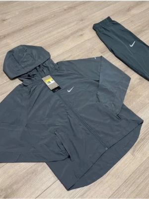 🩶Grå träningsset från Nike med huva🩶 - Snyggt grått träningsset från Nike med jacka och byxor. Jackan har huva, dragkedja och två sidofickor samt Nike-logga på bröstet. Byxorna har resår i midjan och vid benslut samt liten Nike-logga. Perfekt för löpning eller gym. Materialet är lätt och snabbtorkande. Passar dig är cirka 160-170 beroende på vikt också. Tar emot rimliga bud. Byxorna är jätte lite mörkare än överdelen fast man mörkar knappast.