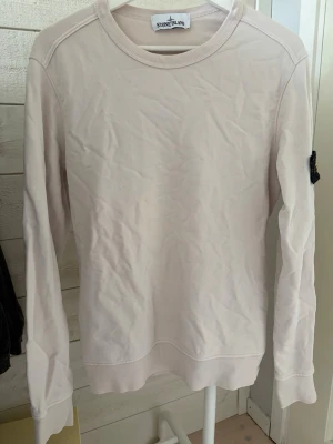  Stone Island beige sweatshirt  - Säljer en stilren ljusbeige sweatshirt från Stone Island med klassisk rund hals och lång ärm. Tröjan har den ikoniska svarta logotypen på vänster ärm och är tillverkad i mjukt bomullsmaterial. Perfekt för en clean och avslappnad look.