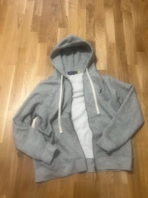 Grå zip hoodie från Polo Ralph Lauren - Snygg grå hoodie från Polo Ralph Lauren med dragkedja och vit dragsko i huvan. Klassisk design med liten broderad logga på bröstet. Tillverkad i mjukt sweatshirtmaterial som är perfekt för chill dagar. Ribbade muddar vid ärmar och nederkant.