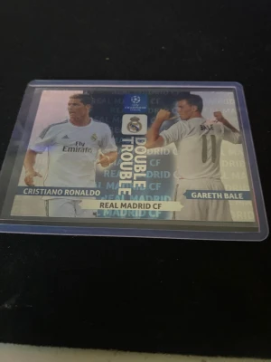 Double Trouble fotbollskort Real Madrid - Säljer ett samlarkort från Panini Adrenalyn XL med Cristiano Ronaldo och Gareth Bale i Real Madrid CF. Kortet har texten 'Double Trouble' och visar båda spelarna i vita matchtröjor. Perfekt för dig som gillar fotboll och samlar på unika kort.