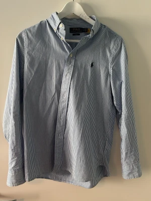Blårandig skjorta från Polo Ralph Lauren - Klassisk blårandig skjorta från Polo Ralph Lauren med vit knappslå och broderad logga på bröstet. Skjortan har lång ärm, button-down krage och är tillverkad i mjuk bomull. Perfekt för en stilren och avslappnad look.