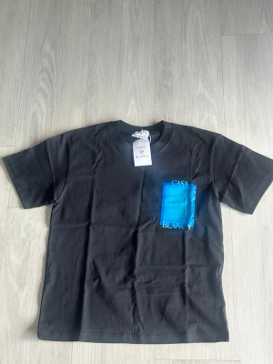 Svart t-shirt med blått tryck - Snygg svart t-shirt med ett stort blått grafiskt tryck och texten 'CASA BLANCA' på bröstet. Klassisk passform och rund halsringning. Perfekt för dig som vill ha en stilren men ändå unik look.