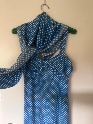 Blå prickig klänning med rosett - Säljer en blå klänning med vita prickar och en stor rosett framtill. Klänningen har smala axelband och är i en lätt, luftig modell som passar perfekt till sommaren. Materialet känns som bomull eller polyester och mönstret är klassiskt prickigt.